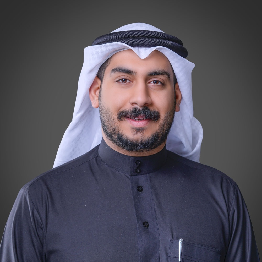 رجل يشبه شخصية أحد عملاء الأرشيف، يرتدي ثوبًا أسود وغترة بيضاء مع عقال داكن، ويبتسم بحرارة على خلفية رمادية داكنة.