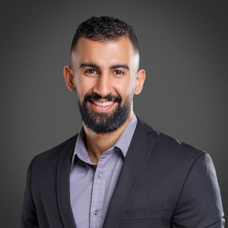 Ahmed AlBossta, a man with a beard, smiles while wearing a black suit jacket and gray shirt against a gray background.