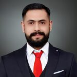 Ahmed AlBossta, a man with dark hair and beard, wears a sharp dark suit complemented by a crisp white shirt and striking red tie, all set against an elegant grey background.