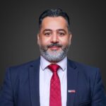 Ahmed AlBossta, a man with a beard, is elegantly dressed in a navy suit and red tie, complete with his name tag. He's striking a pose against a plain dark background.