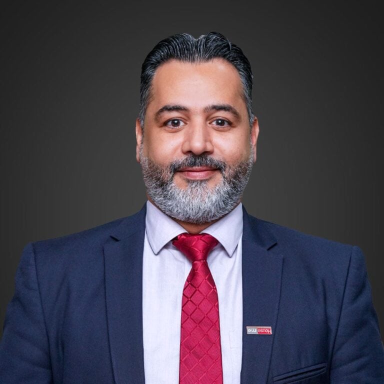 Ahmed AlBossta, a man with a beard, is elegantly dressed in a navy suit and red tie, complete with his name tag. He's striking a pose against a plain dark background.