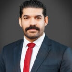 A man with a mustache wearing a dark suit, white shirt, and red tie against a plain gray background.