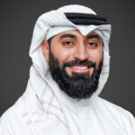 A man with a beard, identified as Jaffer Mirza, is wearing traditional white Middle Eastern attire and a headscarf, smiling against a dark background.