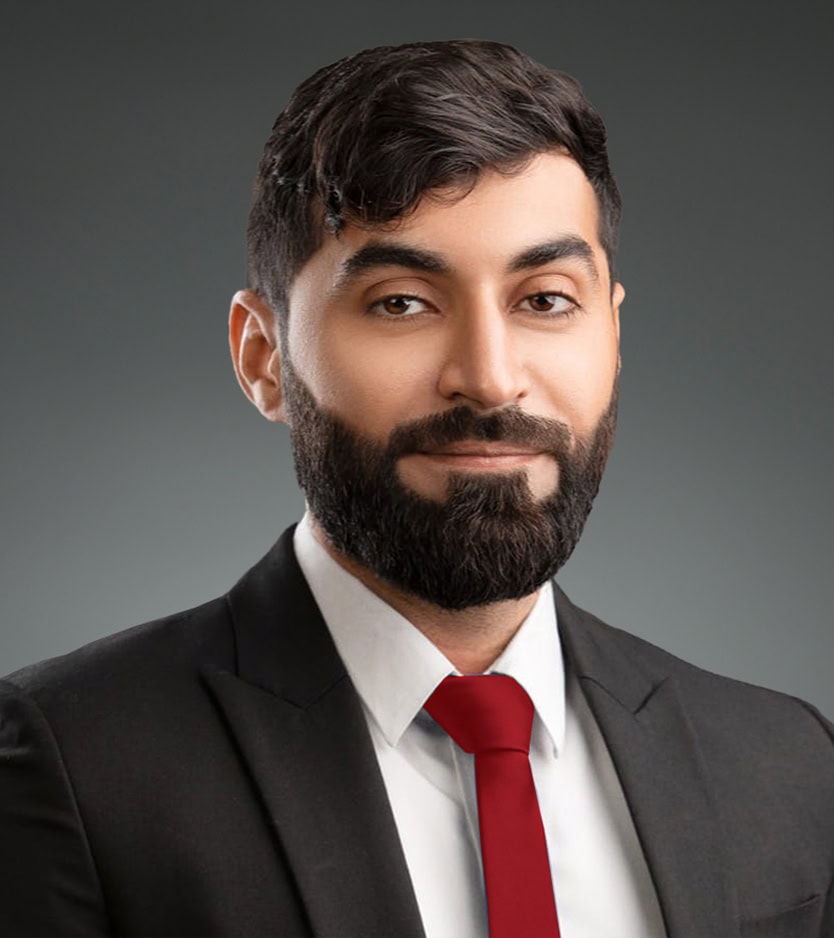Portrait of Jaffer Mirza with a beard, wearing a black suit, white shirt, and red tie, looking at the camera with a light smile. The background is solid gray.