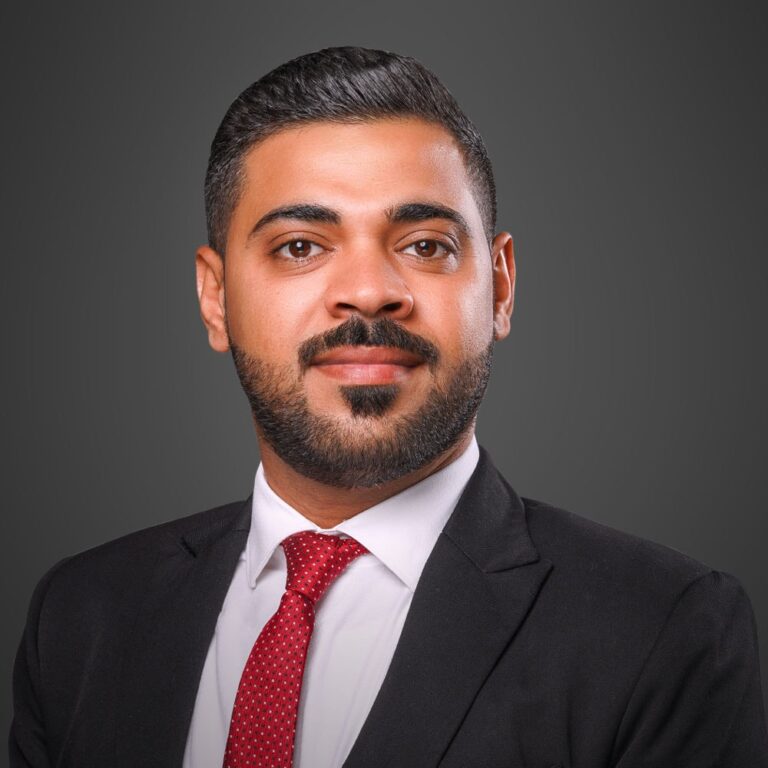 Ahmed AlBossta, dressed in a sharp suit with a red tie and sporting a well-groomed beard, poses confidently against a gray background.