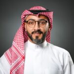 Ahmed AlBossta stands gracefully, wearing glasses, a red and white checkered keffiyeh, and a pristine white thobe against a subtle gray background.