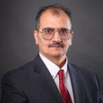 Ahmed AlBossta, a man with glasses and a mustache, wears a suit paired with a red tie, set against a gray background.