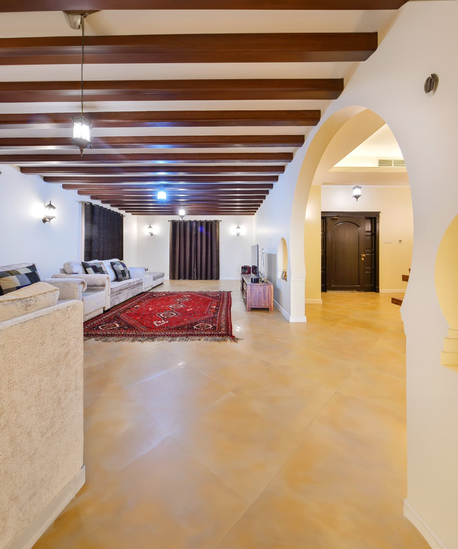 Spacious living room in this 3BR Villa at Al Areen features a beige tile floor, wooden ceiling beams, arches, a red patterned rug, beige sofas, a TV on a wooden stand, and dark curtains—a perfect complement to the big garden just outside.