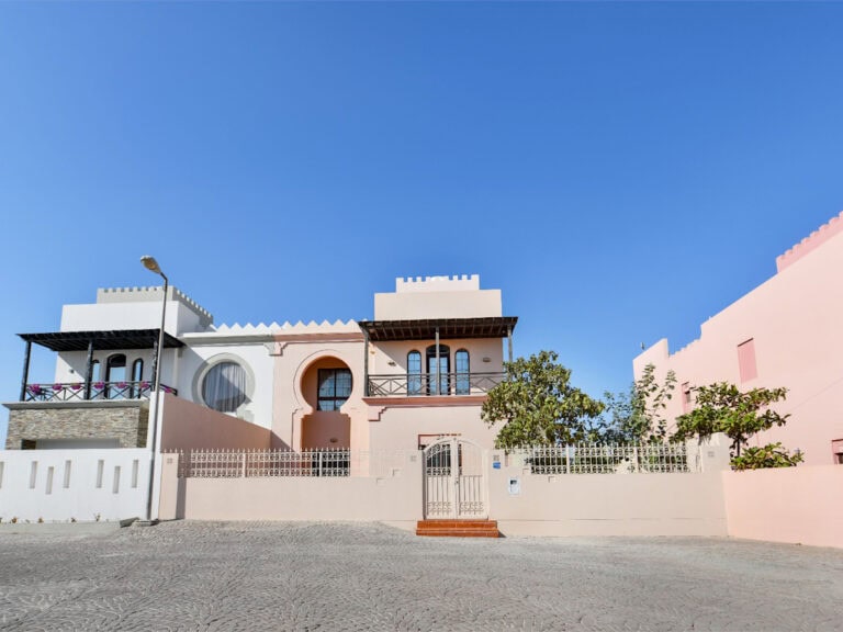 A large, light pink 3BR villa for sale with unique arched windows and a gated entrance under a clear blue sky, flanked by similar buildings on either side. Located in Al Areen, it boasts a big garden perfect for outdoor gatherings.