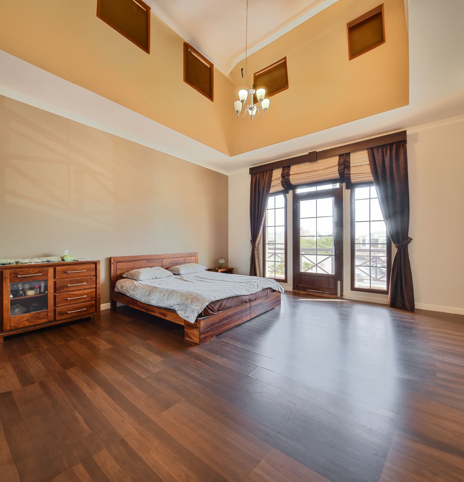 A spacious bedroom with a high ceiling, wooden floor, a bed with white and brown bedding, a wooden dresser, and large windows with curtains allowing natural light to fill the room. This serene space is part of the 3BR Villa in Al Areen, complete with a big garden for outdoor relaxation.