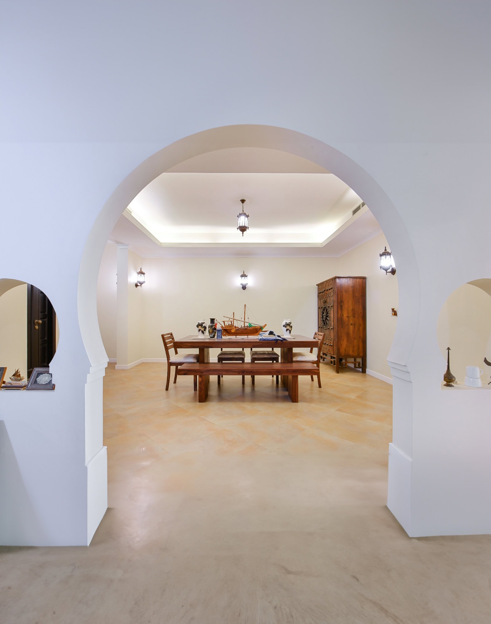 A spacious room in the 3BR Villa at Al Areen features white walls and a wooden dining table with chairs under drop lantern lights. The table, adorned with a model ship, is surrounded by art pieces set in arch-framed walls. This villa also boasts a big garden for ultimate relaxation.