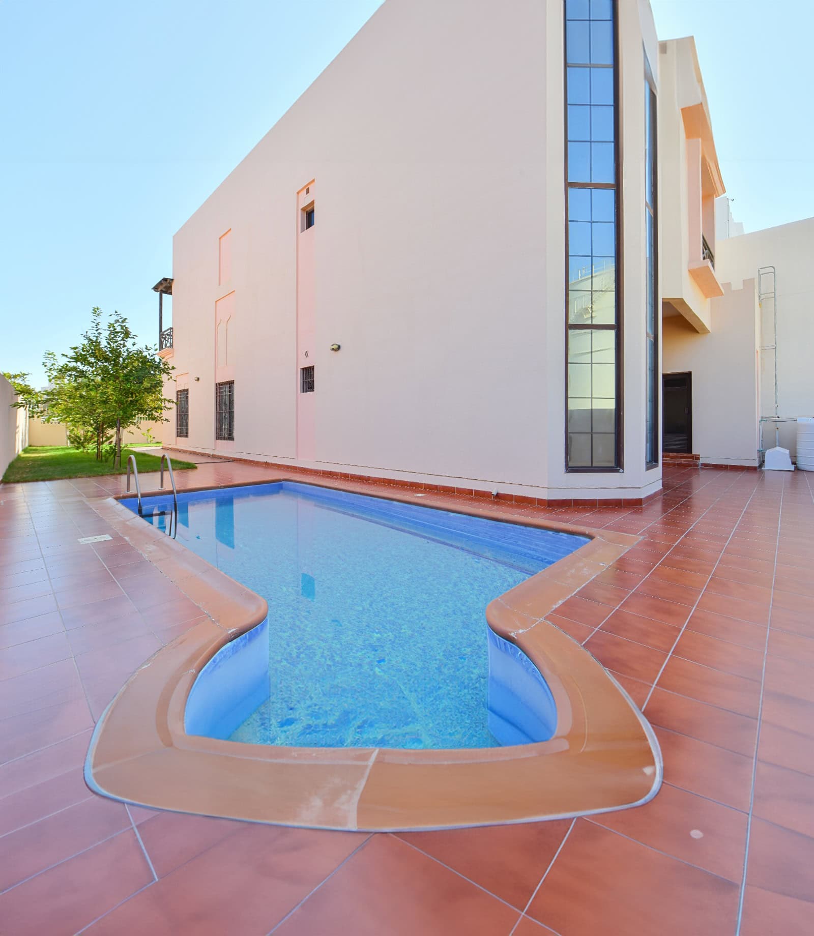 A narrow, curved swimming pool with red-tiled flooring lies in the backyard of a modern 3BR villa. The pool is surrounded by a beige border, adjacent to the stylish two-story building, with a tree standing proudly in the background amidst the big garden.