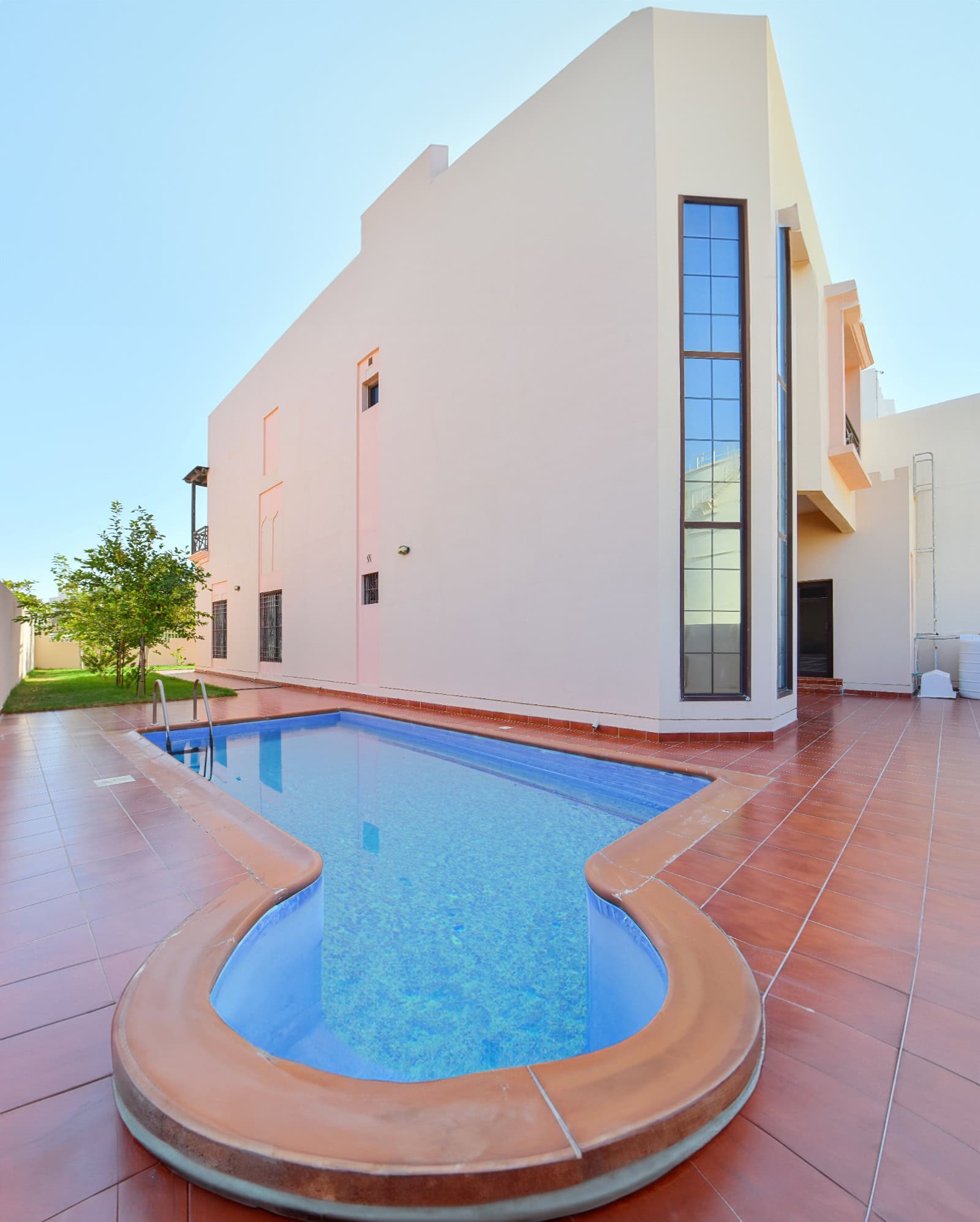 A modern 3BR villa in Al Areen boasts a two-story white structure with tall windows, a big garden, and a narrow, kidney-shaped outdoor pool lined with reddish tiles in the backyard.