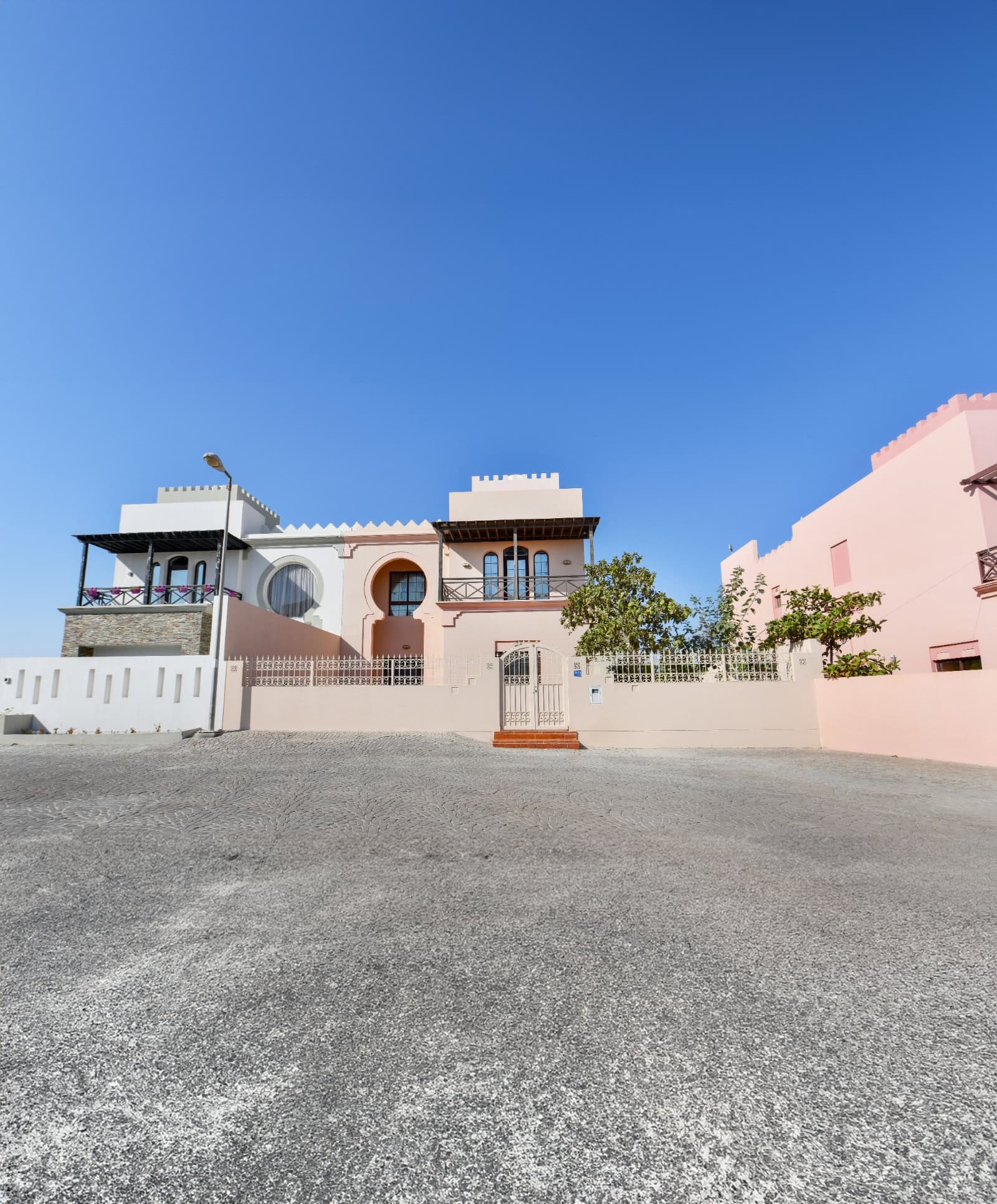 A pink and white building with a castle-like design stands against a clear blue sky, featuring arched windows, decorative balconies, and a gated entrance. The 3BR Villa in Al Areen is complemented by its expansive big garden, adding to the fairytale charm.