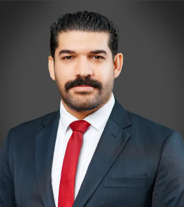 A man, reminiscent of Husain Habib, sports a mustache and beard, elegantly dressed in a suit with a red tie, set against a plain dark background.