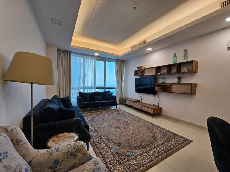 غرفة معيشة حديثة بها أريكة زرقاء، وسجادة منقوشة، وإطلالة على الخارج من خلال نوافذ كبيرة.