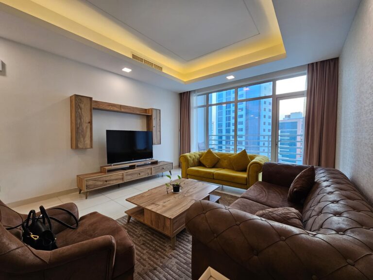 غرفة معيشة حديثة بها أريكة جلدية بنية اللون، ومقعدين أصفر اللون، وأثاث خشبي، وتلفزيون، وإطلالة على المدينة من خلال النافذة الكبيرة.