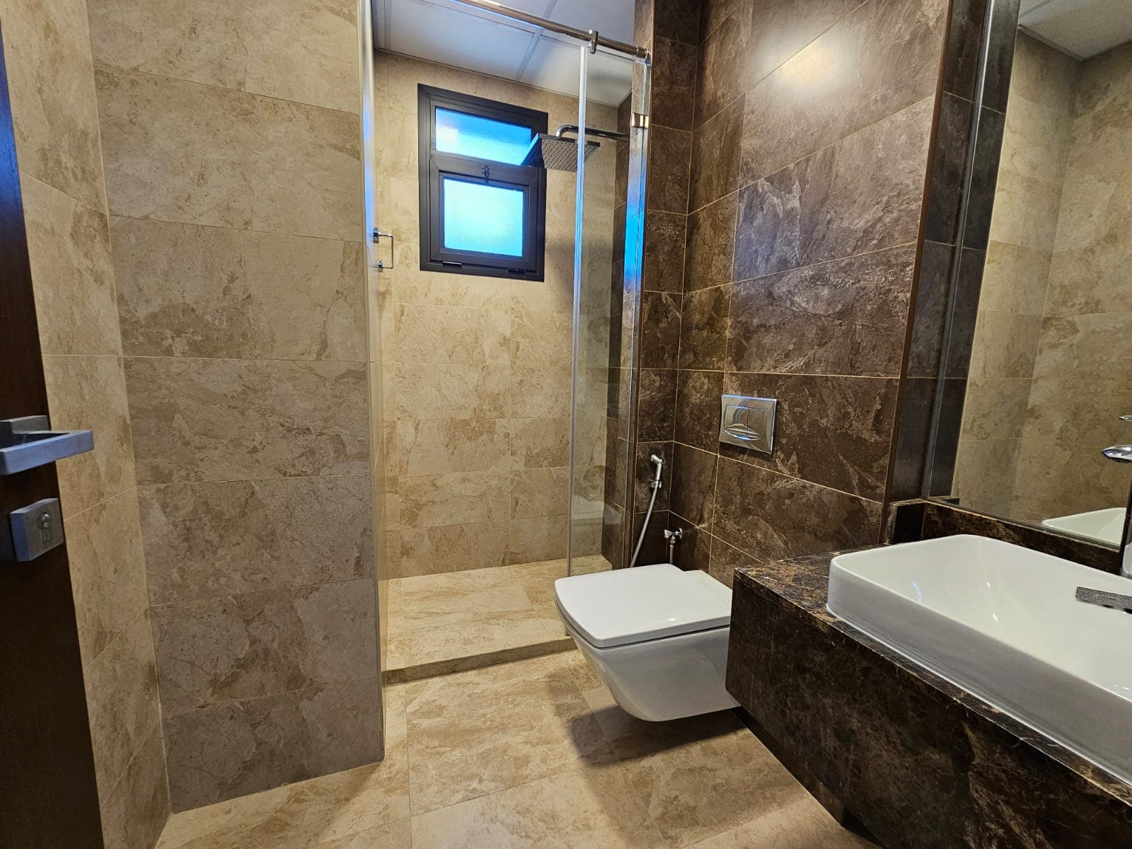 Modern luxury apartment bathroom interior in the Juffair area with glass shower enclosure, wall-mounted toilet, and dark marble accents.