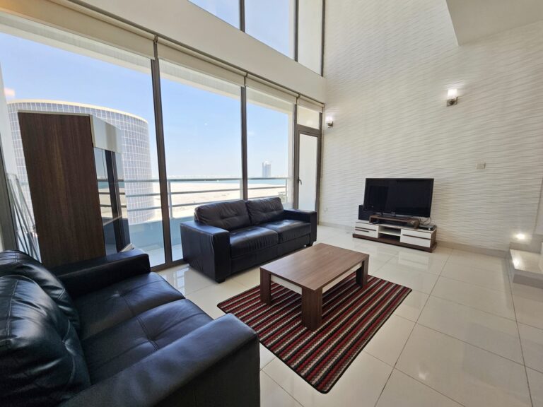 A bright, modern 3-bedroom Juffair flat with large windows offering a view of the skyline, furnished with black sofas, a wooden coffee table, and a television set.
