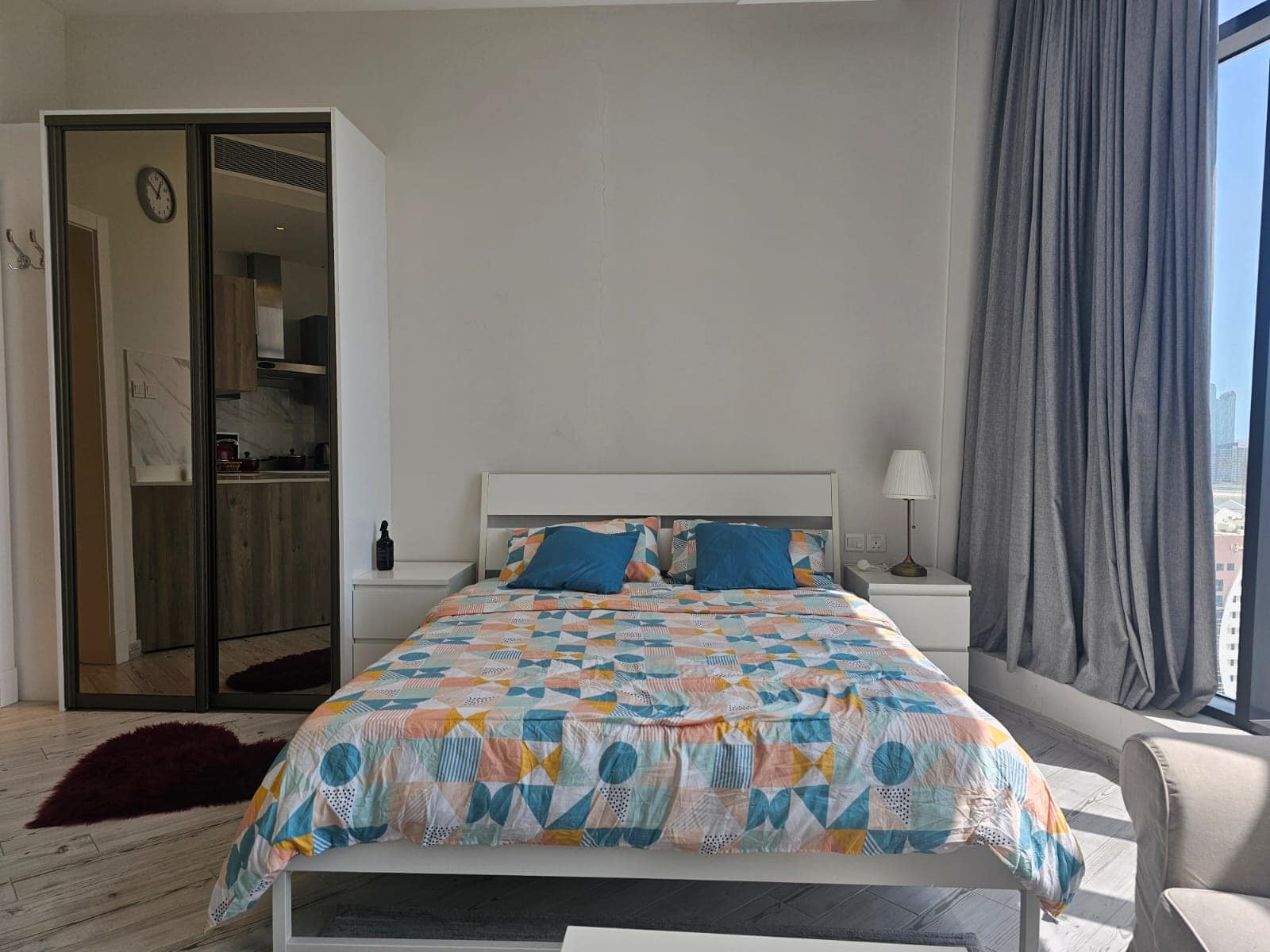 Luxury apartment for rent; modern bedroom with a patterned quilt, floor-to-ceiling window, and a view of the city skyline in the Seef area.