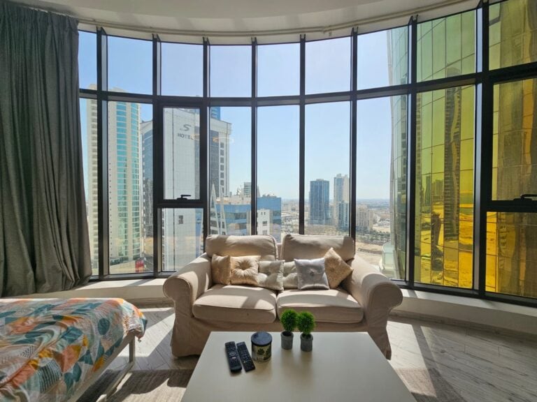A modern luxury apartment living room with a curved window wall offering a panoramic view of a city skyline, available for rent in the Seef area.