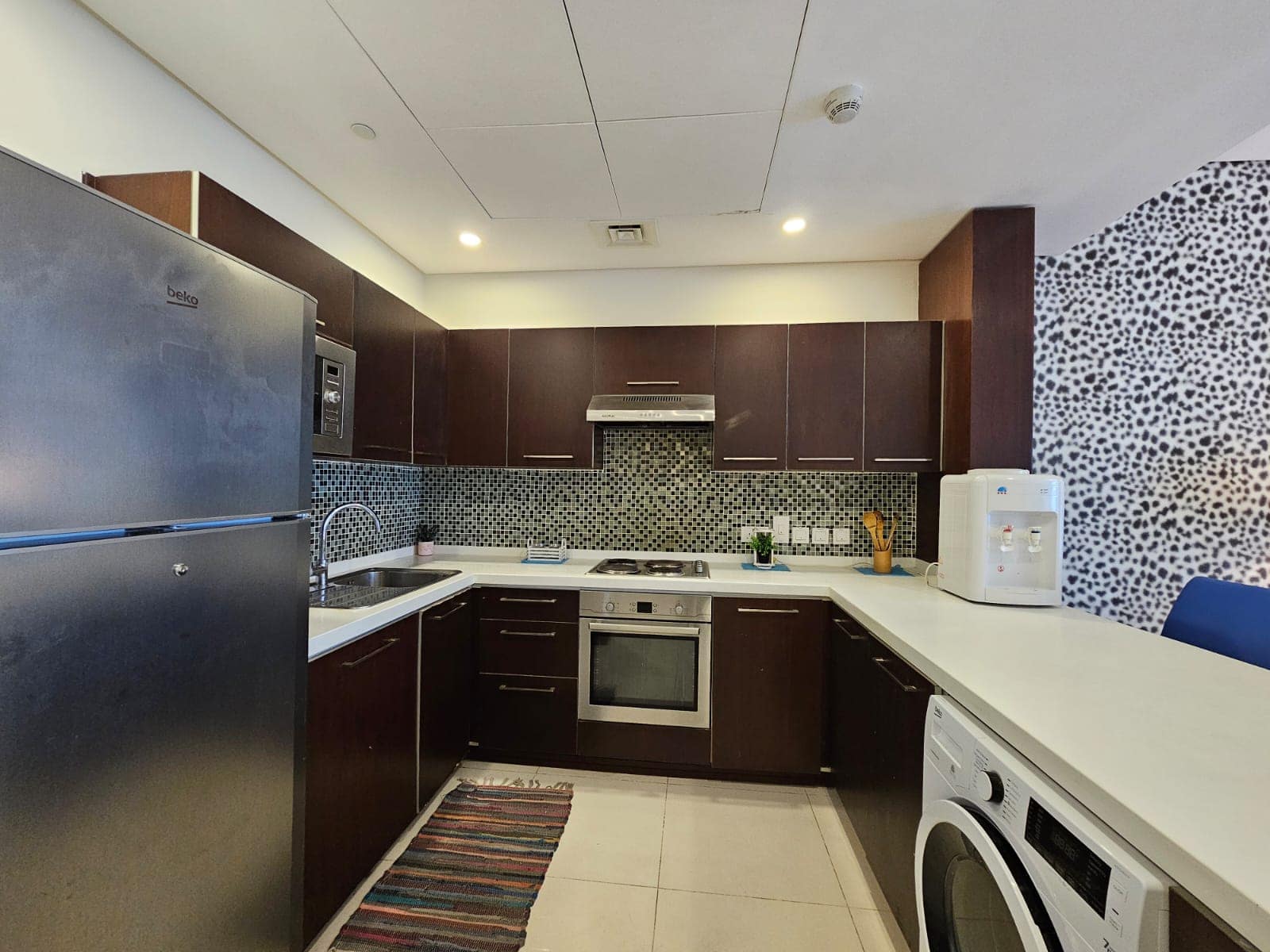 A modern kitchen in an apartment for rent in Juffair with stainless steel appliances, dark wood cabinets, and a decorative tiled backsplash.