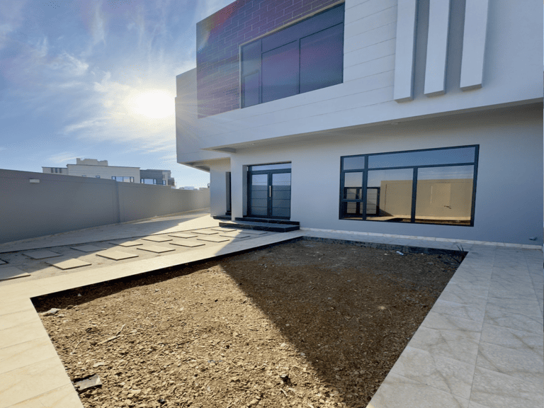 Elegant villa facade with large windows and a small courtyard area under a clear blue sky in the Al Hajar area.