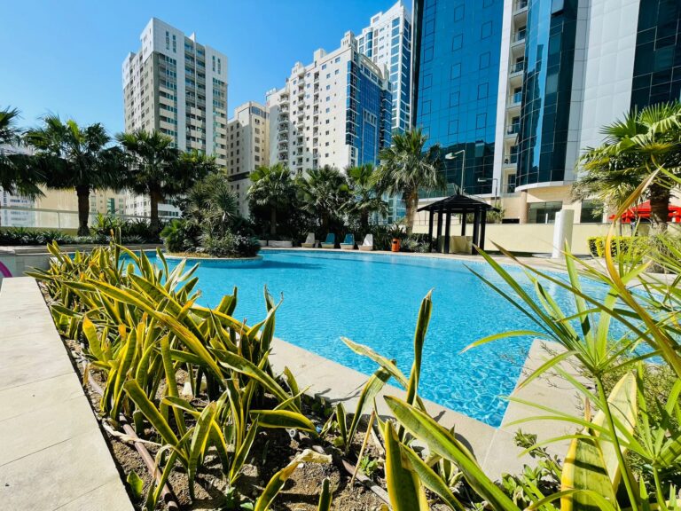 Outdoor swimming pool surrounded by high-rise buildings and landscaped with green plants under a clear blue sky, part of a luxury 2BR apartment for rent in Juffair.