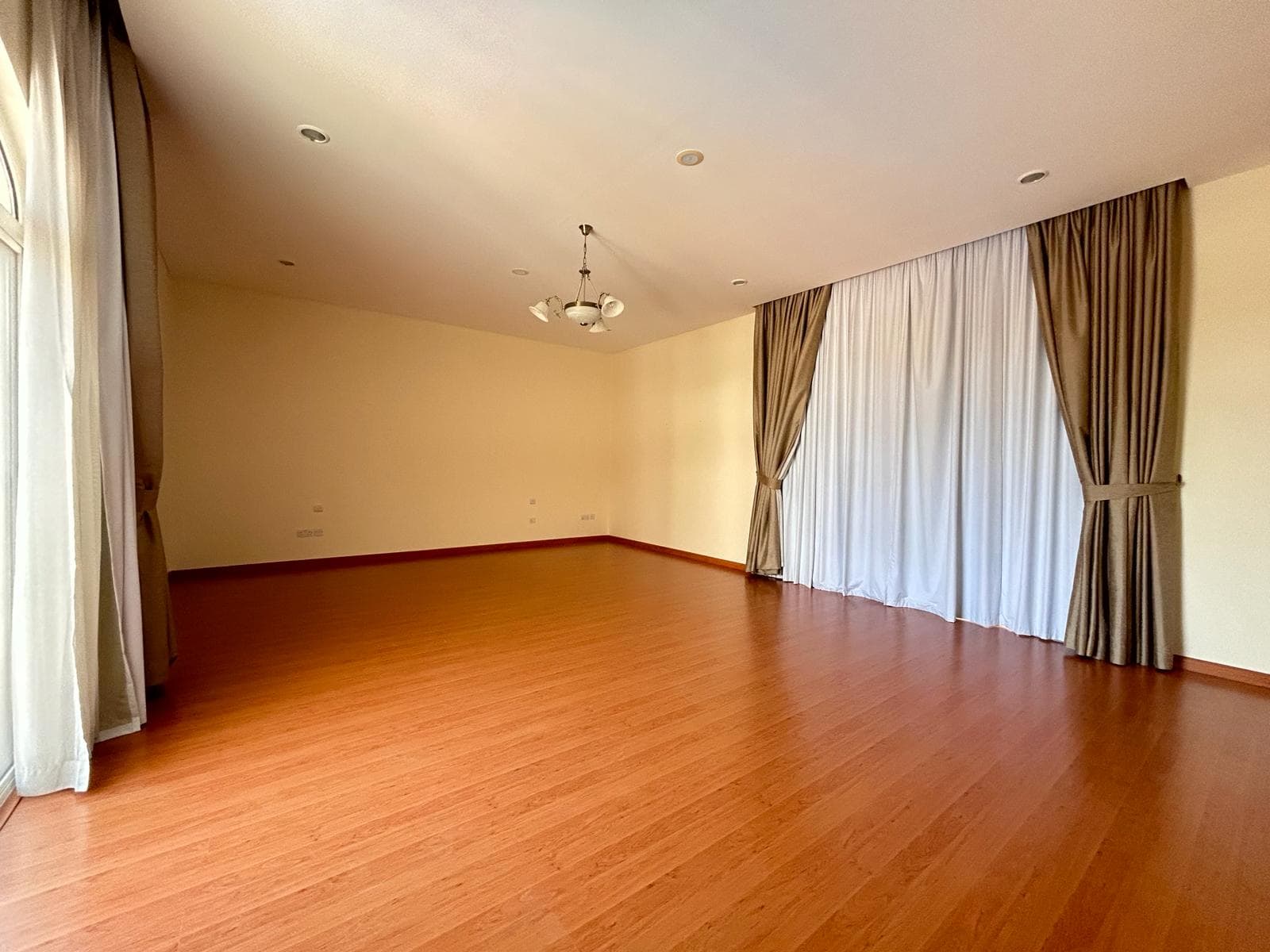 Empty room with wooden flooring and large windows covered by curtains in a villa for rent, located in the Compound Hamala, Hamala area.