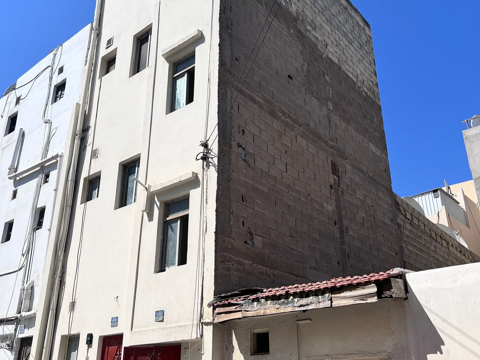 A bare side wall of the Salmaniya building with no windows, adjacent to a structure with a corrugated metal awning, under a clear blue sky.