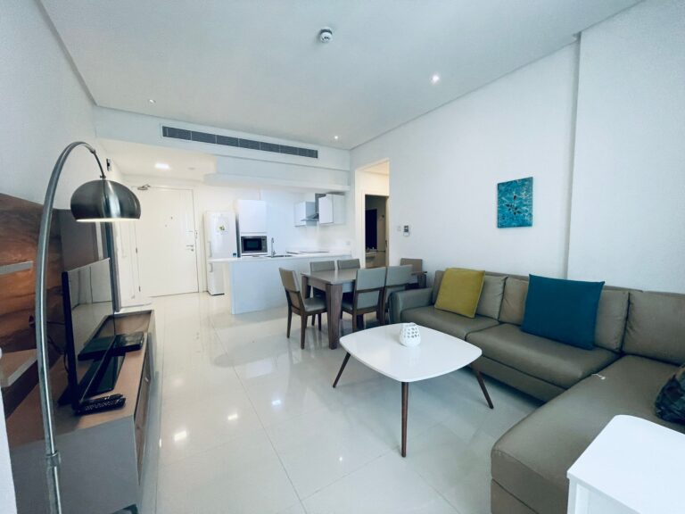 غرفة معيشة حديثة مع مطبخ مفتوح وأريكة مقطعية ومنطقة لتناول الطعام وديكور بسيط.
