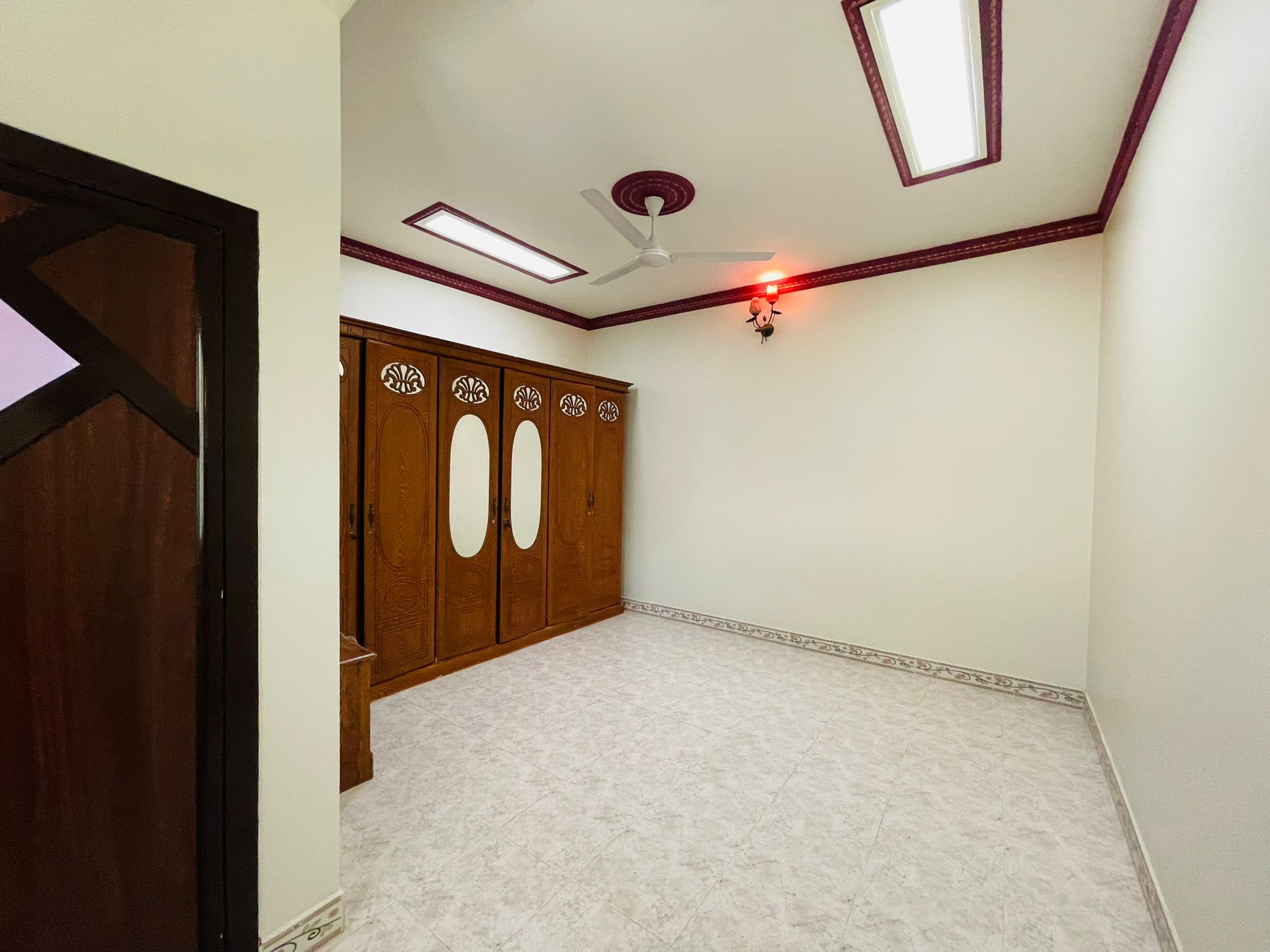 An empty 2BR Villa in Ras Rumman with white tiled flooring, off-white walls with decorative borders, a ceiling fan, and a wooden double door closet.