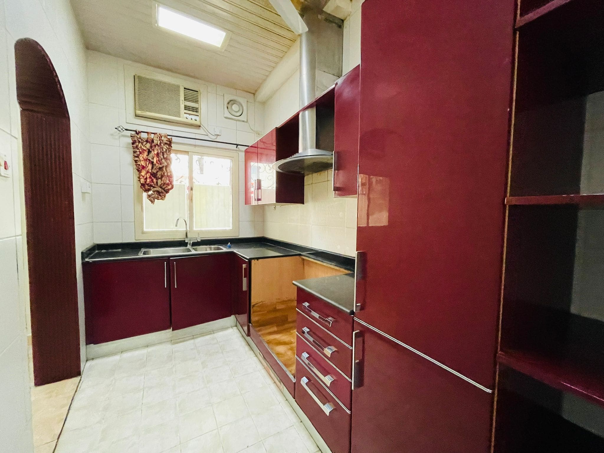 A compact kitchen in a 2BR Villa with maroon cabinets, white tiled flooring, and an air conditioning unit above the window.