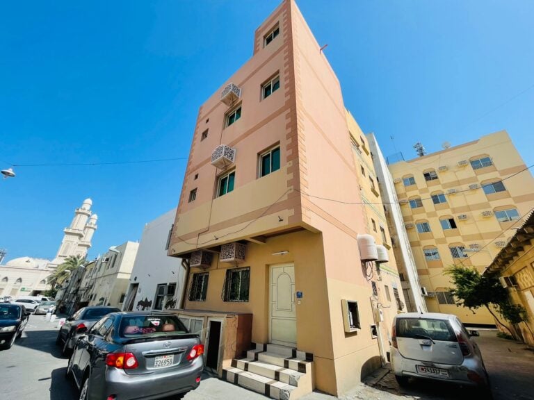 A peach-colored corner building on a sunny street with parked cars and a minaret in the background, now serves as a semi-furnished 2BR villa in Ras Rumman.