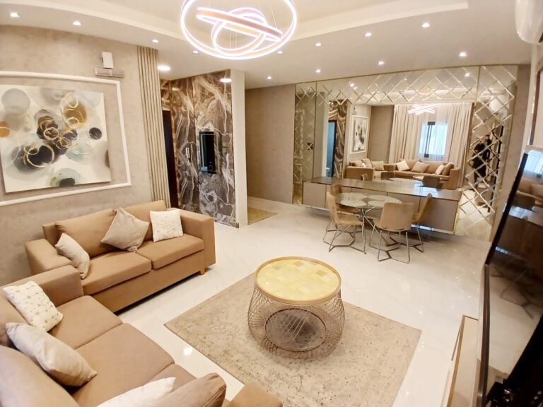 A modern living room interior with a beige sofa, a circular center table, artwork on the wall, and an adjoining dining area with stylish lighting. Auto Draft