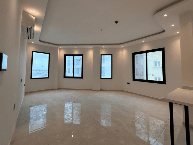 An empty apartment for rent, with white walls, polished tile flooring, and multiple windows allowing natural light in the Janabiyah area.