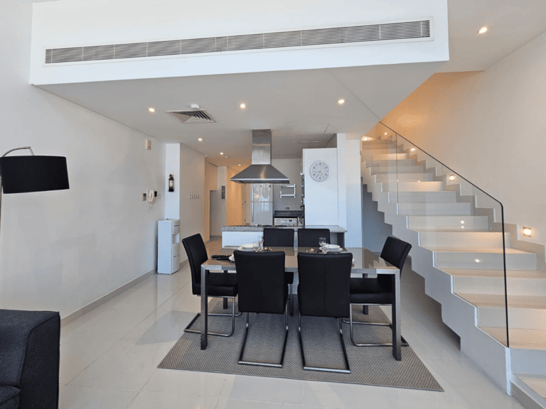 A modern dining room with a staircase leading to the upper level, featuring a glass balustrade and clean, white interior design.