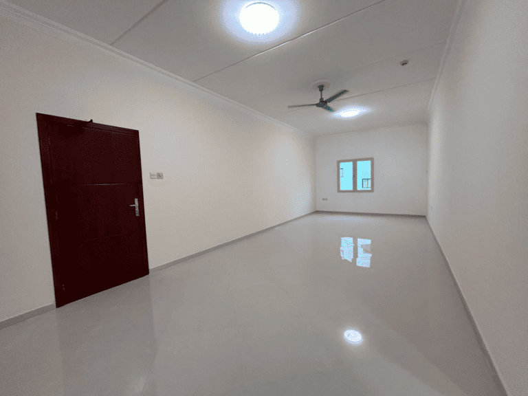 An empty apartment in Janabiyah Area with glossy tiled flooring, white walls, a wooden door on the left, and a ceiling fan for rent.