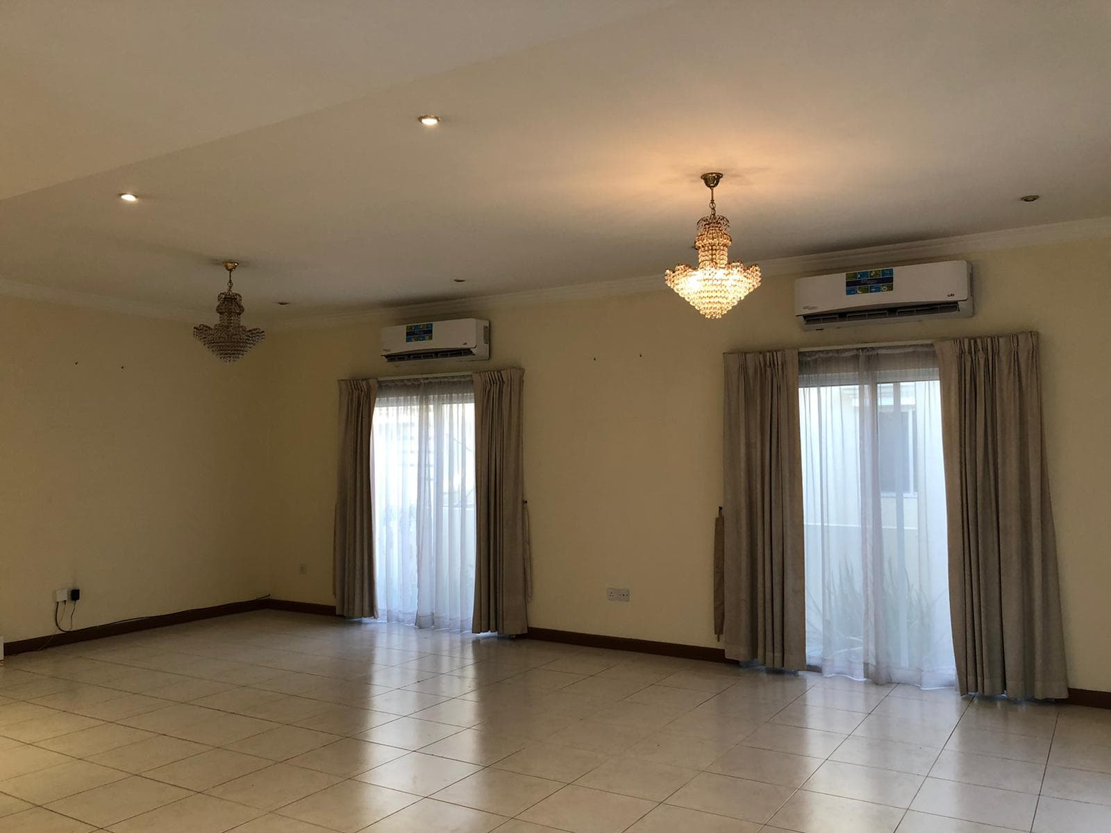 Empty villa living room with tiled floor, two chandeliers, air conditioner, and curtains on windows.