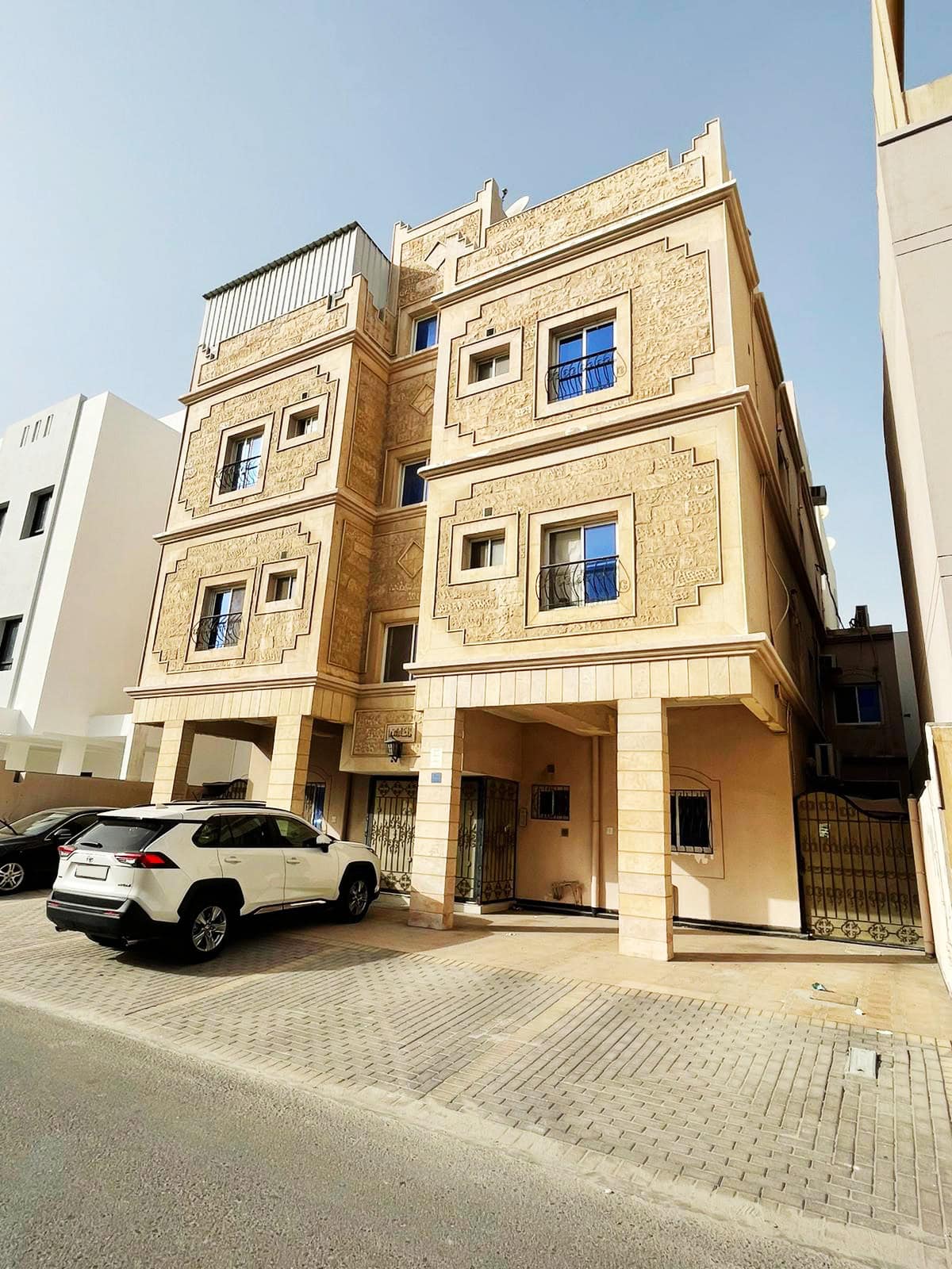 A modern three-story building for sale with ornate beige facades and blue window frames, parked cars nearby, under a clear sky in the New Hidd Area.