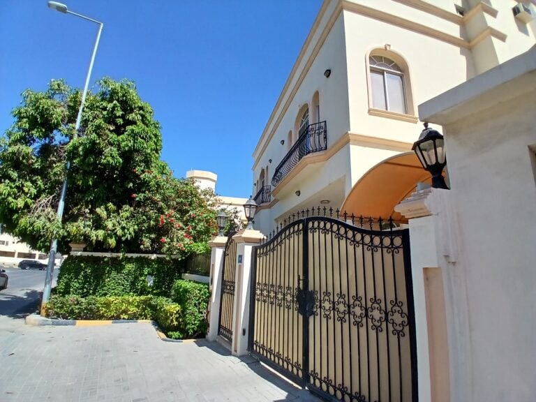 A sunny street view showcasing a large beige house with balconies and an auto draft wrought iron gate surrounded by greenery.