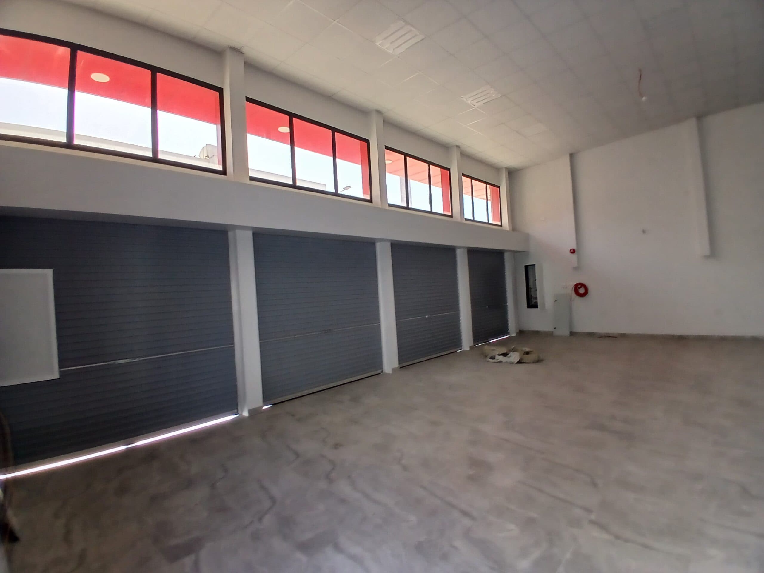 Interior of an empty industrial warehouse with grey flooring and several closed metallic roller shutter doors under a row of high windows with auto draft tint.