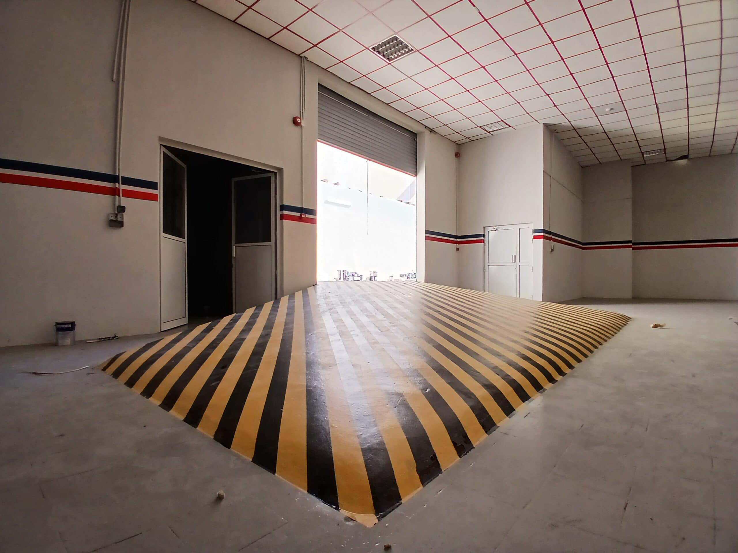 Empty industrial warehouse interior with a striped ramp leading to a higher level, white walls with red and blue stripes, and a large auto draft roll-up door.