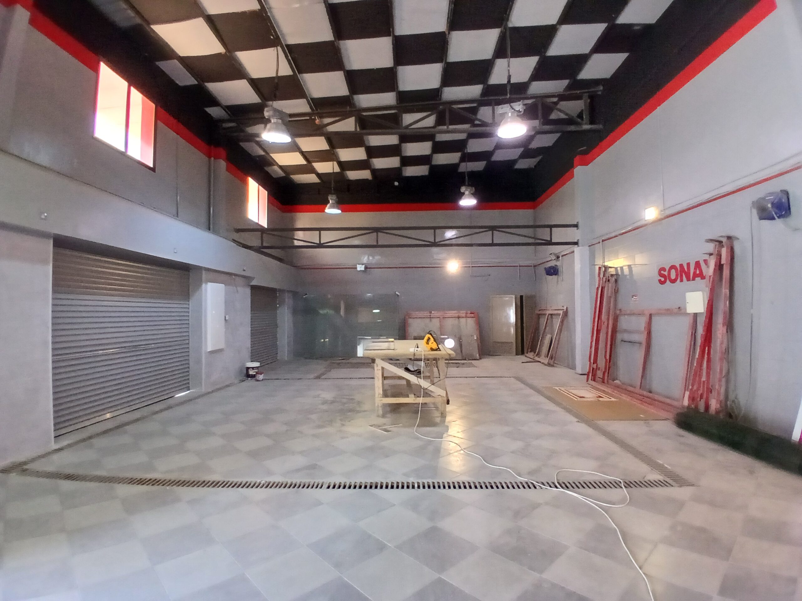 Semi-fitted industrial garage with checkered floor, black and white ceiling, red accents, and metal roll-up doors. A table with tools is in the center.