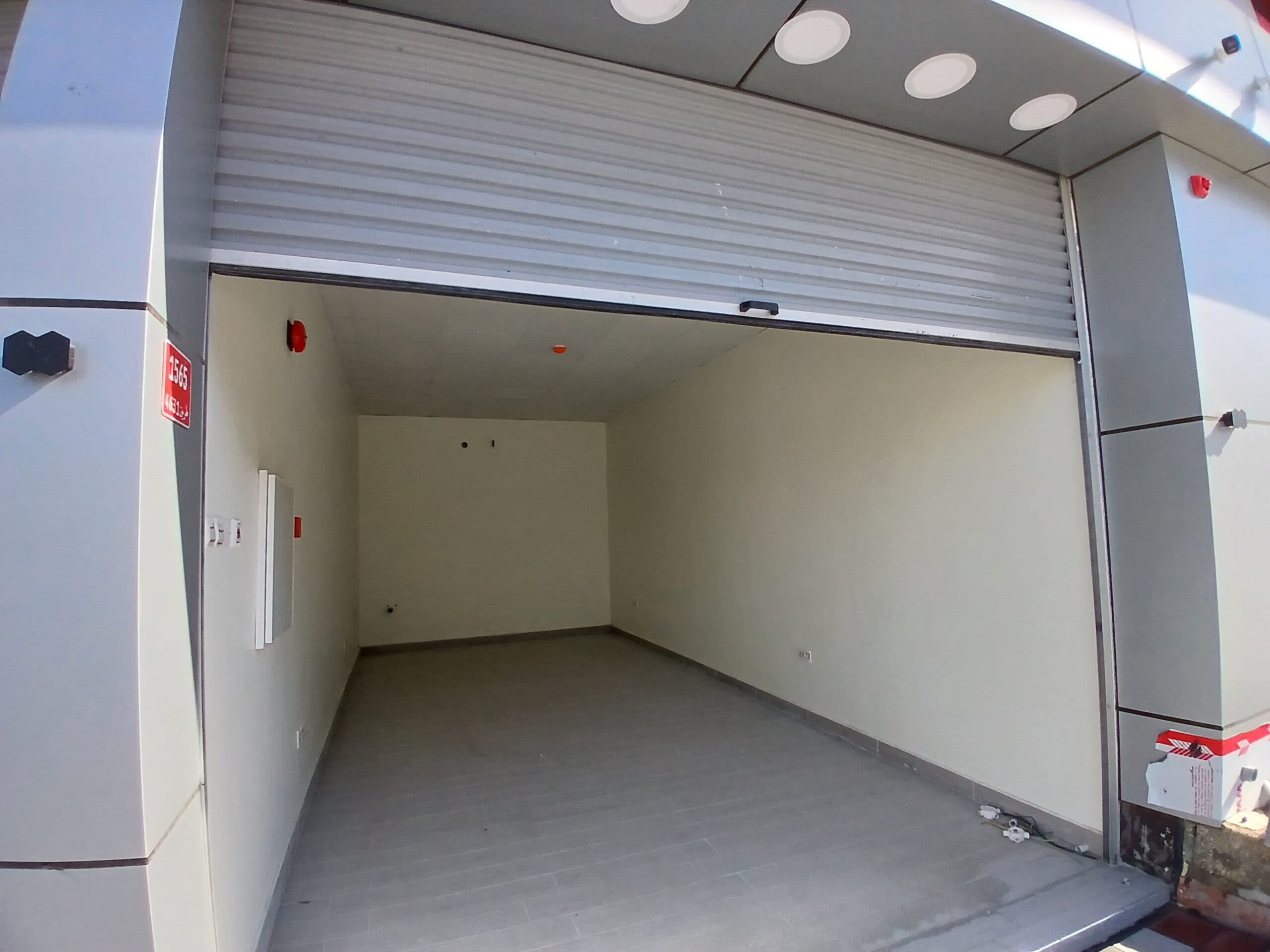 An empty commercial vehicle space with a rolled-up white shutter door and a gray floor; lighting is installed on the ceiling.