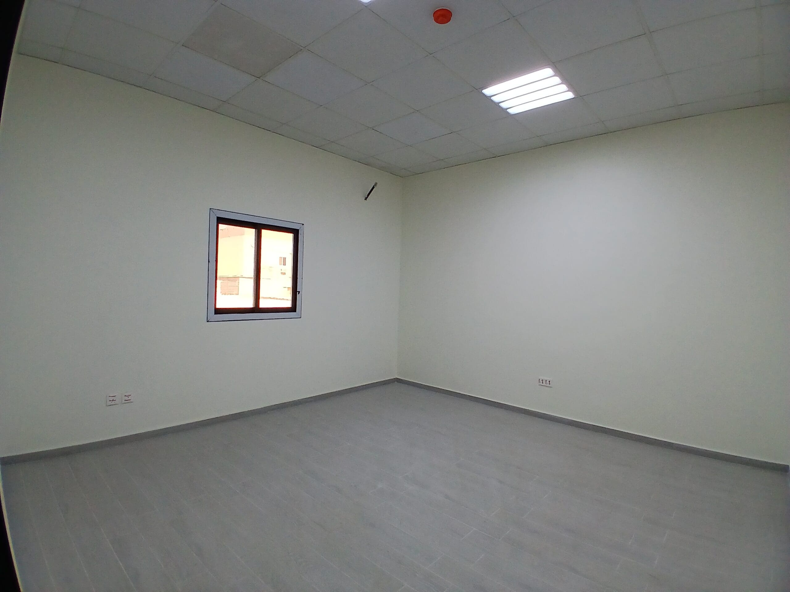غرفة فارغة ذات جدران بيضاء، ونافذة واحدة، وأرضية من البلاط الرمادي.