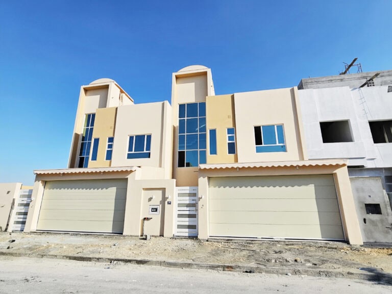 Two luxurious duplex villas in Hamala under a clear blue sky, featuring beige exteriors with large garages and arched windows.