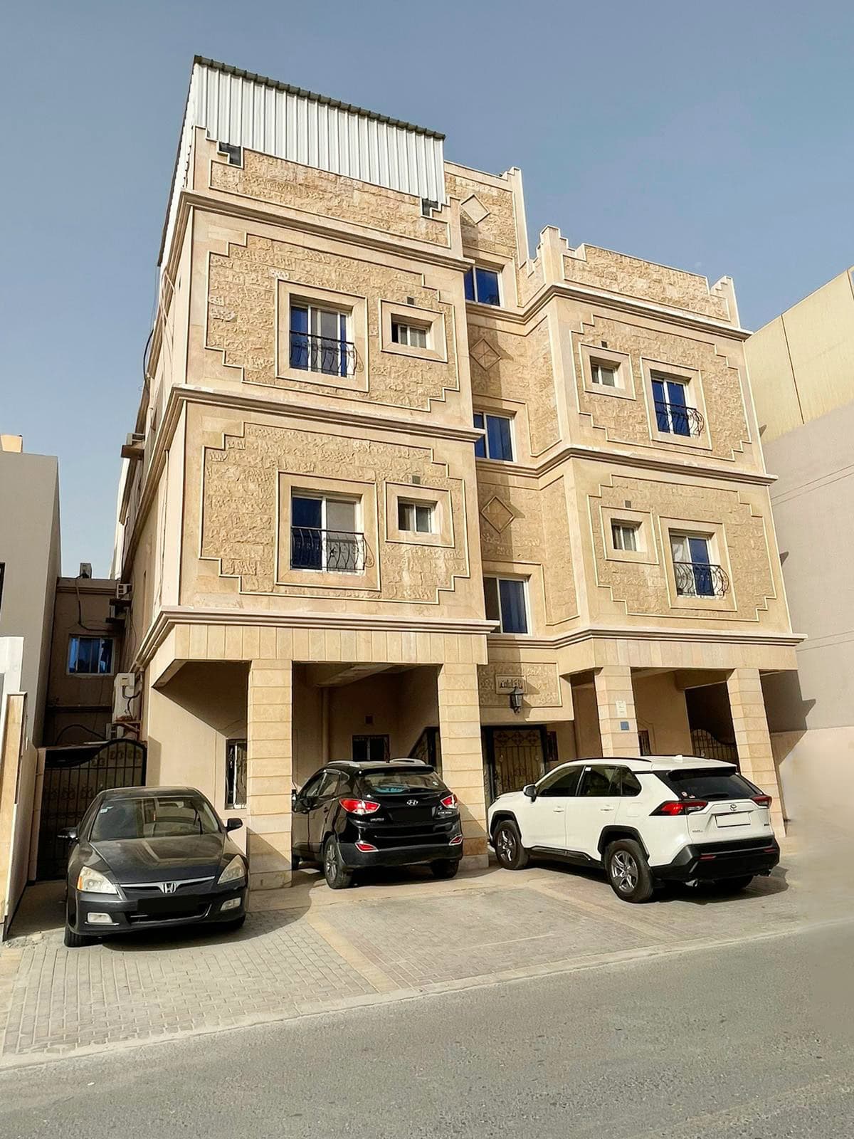 A modern three-story residential building for sale in an urban area, with a textured beige facade and three parked cars in front.