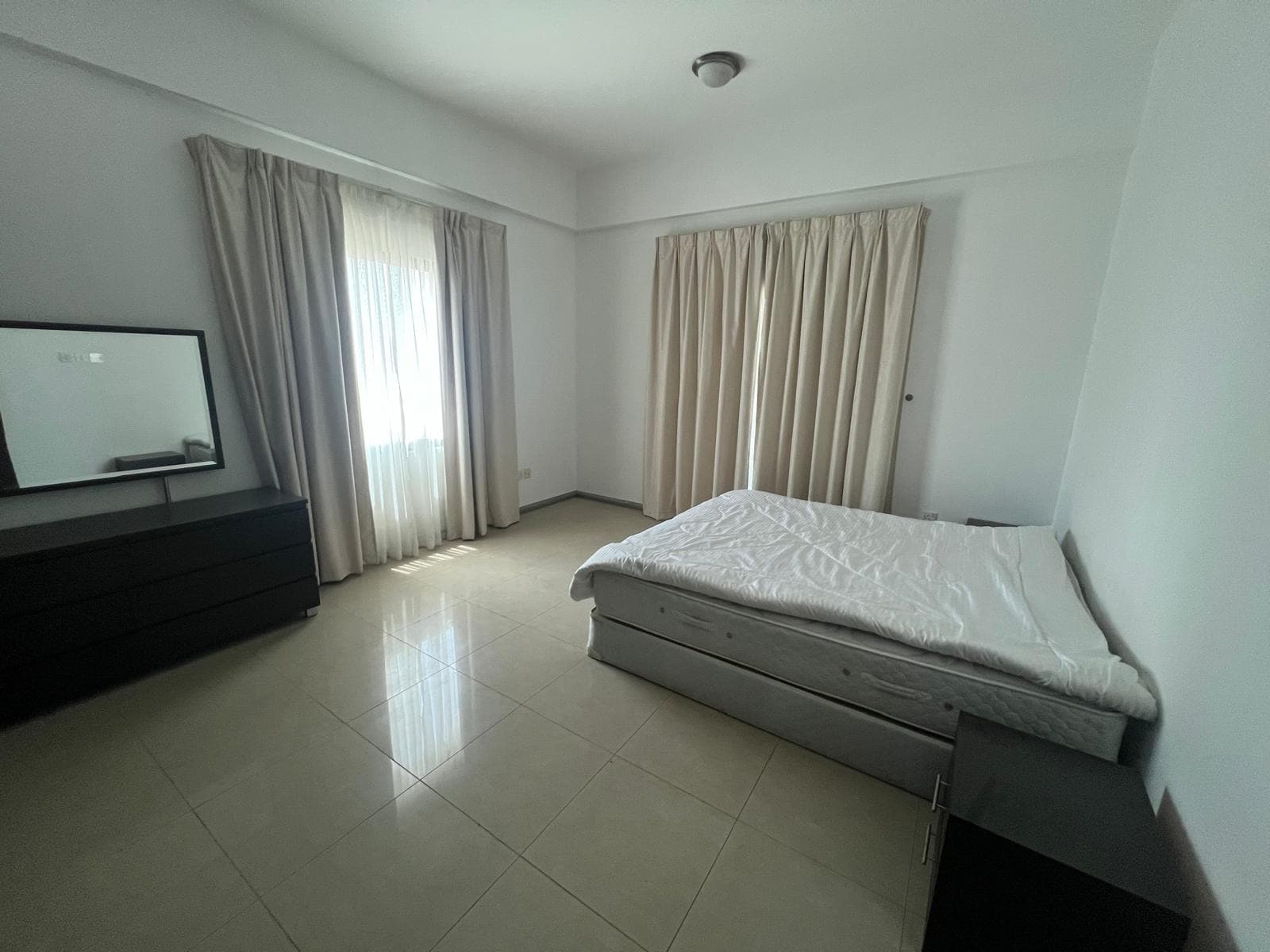 A modern bedroom with a twin bed, dark wood dresser, tv, and large windows with curtains.