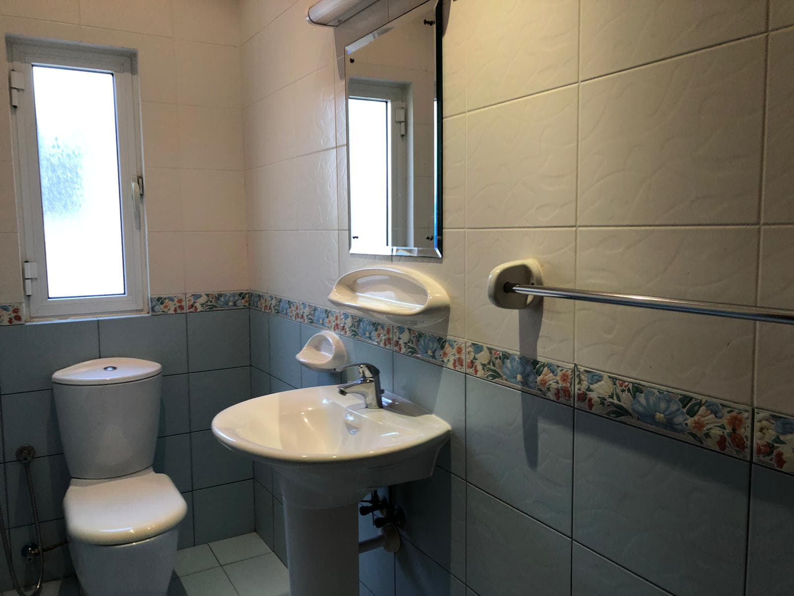 A clean bathroom with a toilet, sink, and mirror in a semi-furnished villa, featuring white and blue tiles and a window on the left.